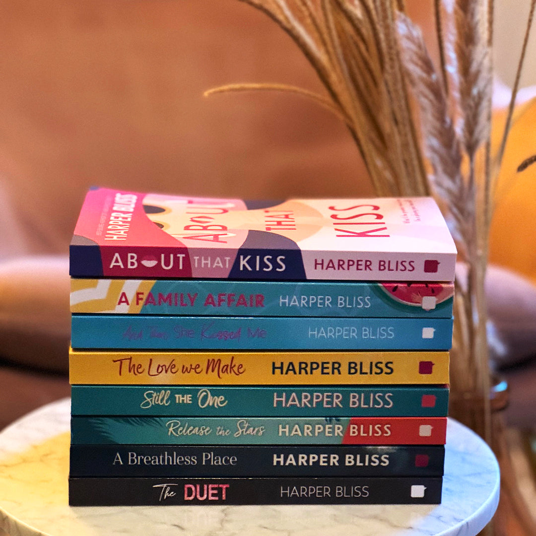 A stack of paperback sapphic romance books on a marble table with some dried flowers in the background. The books in the stack are About That Kiss, A Family Affair, And Then She Kissed Me, The Love We Make, Still the One, Release the Stars, A Breathless Place and The Duet. All books are by Harper Bliss.