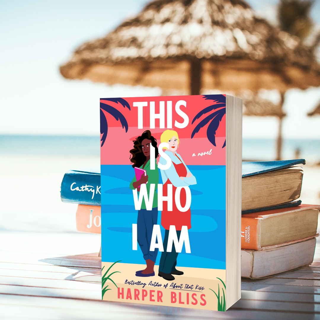 A vibrant image of the book This Is Who I Am by Harper Bliss placed upright on a beach table. The cover features two illustrated women standing back to back—one with dark curly hair holding a book, and the other with short blonde hair in a red apron—set against a pink and blue tropical backdrop with palm trees. The scene includes a blurred beach background with straw umbrellas and a soft-focus stack of books beside the main book, suggesting a relaxing summer reading vibe.